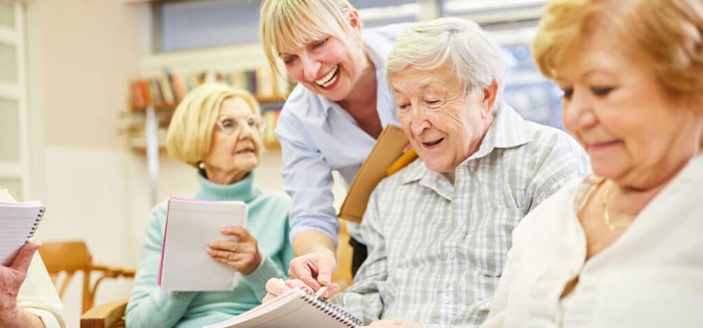 A caregiver helping a group of elderly people write things down in a notebook.